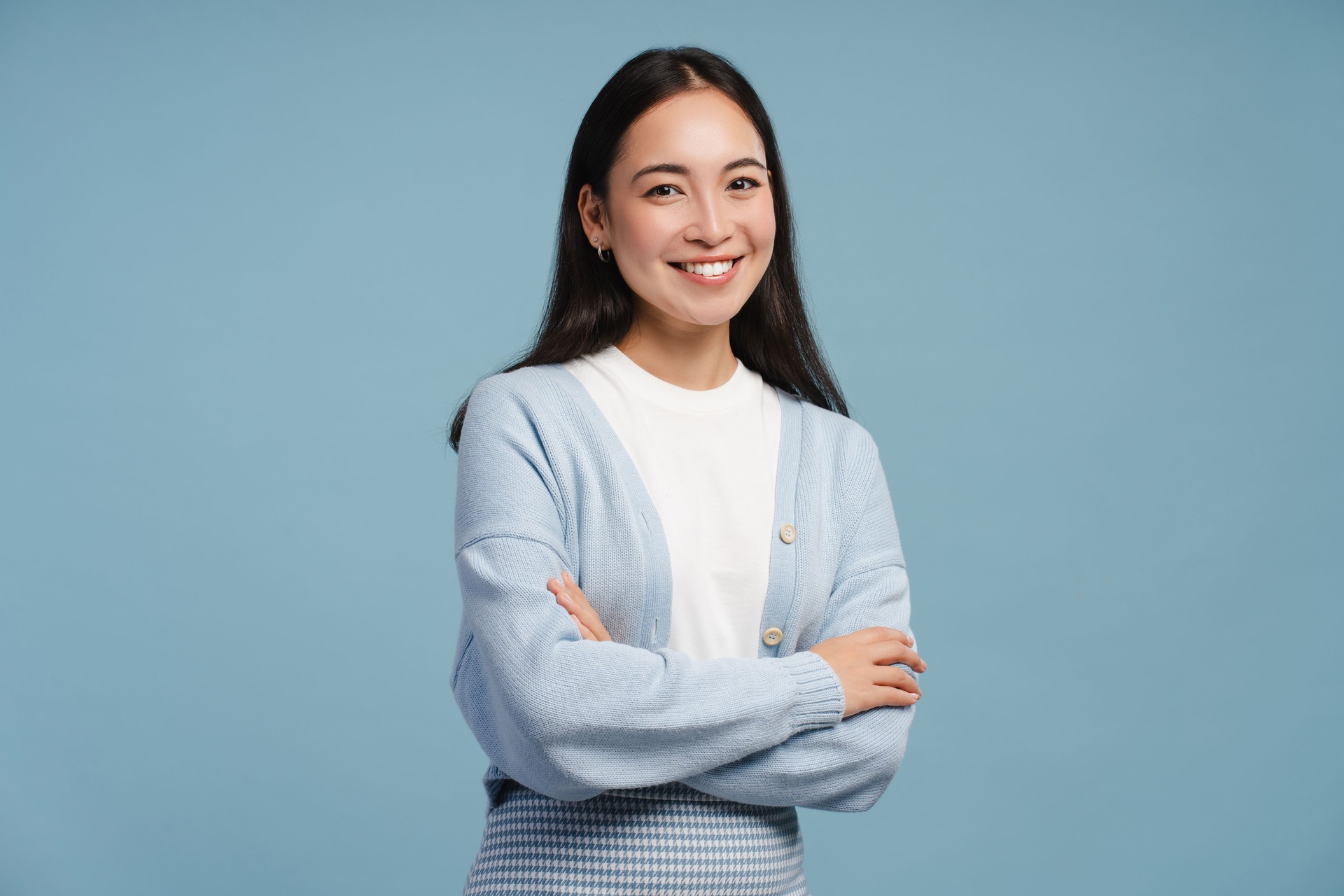 Smiling young Asian woman with crossed arms, looking at camera, standing isolated on blue background